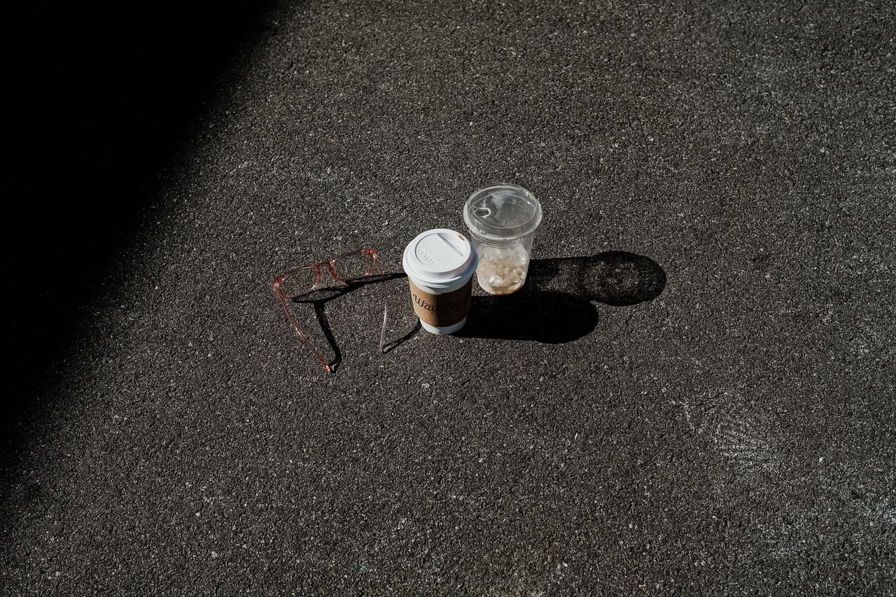 Disposable coffee and plastic cups with eyeglasses on asphalt in sunlight.